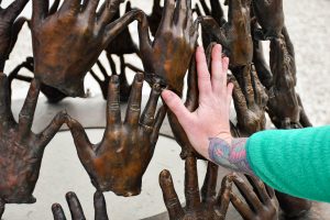 A woman puts her hand up to a metal hand on a sculpture made of many hands.